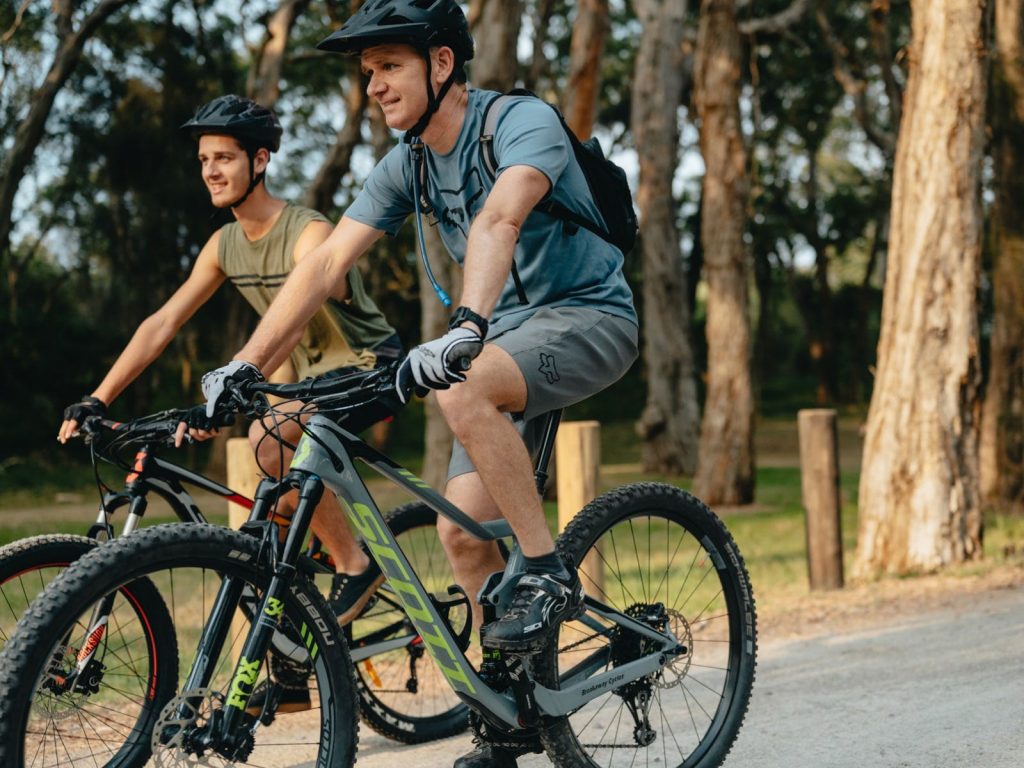Two men riding past on push bikes while wearing helmets.