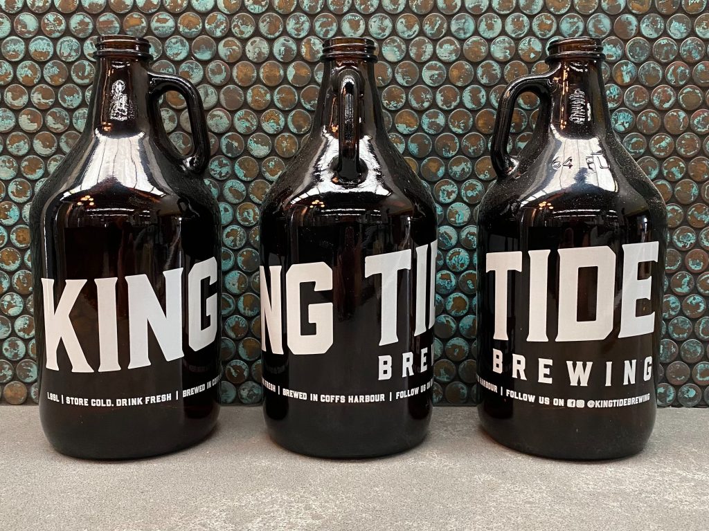 Picture of three growler beer bottles with King Tide Brewing logo, set against copper tile backdrop.