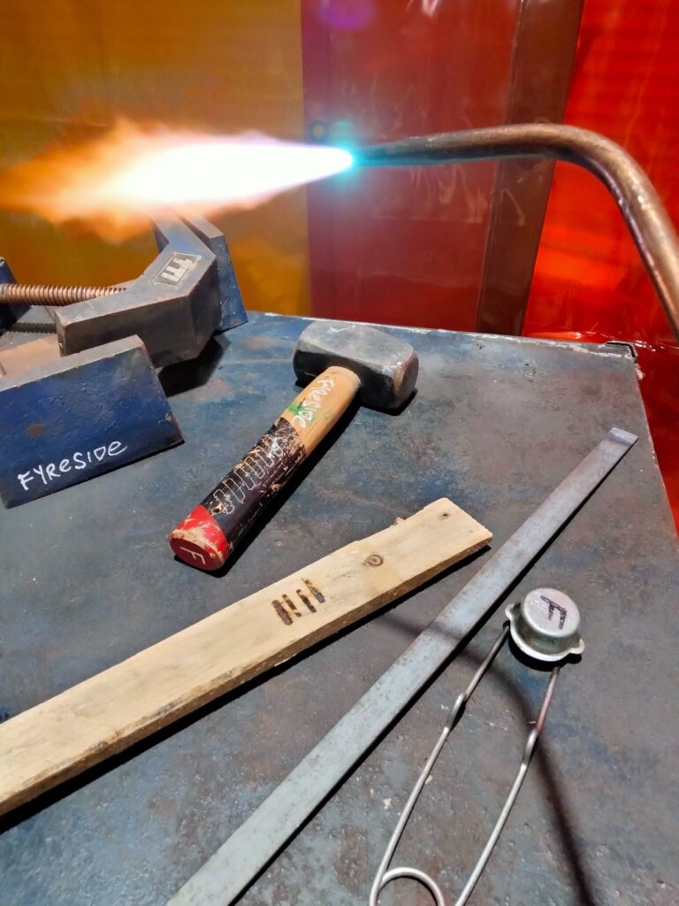 A metal work bench with a vice, hammer, and a flaming welding torch